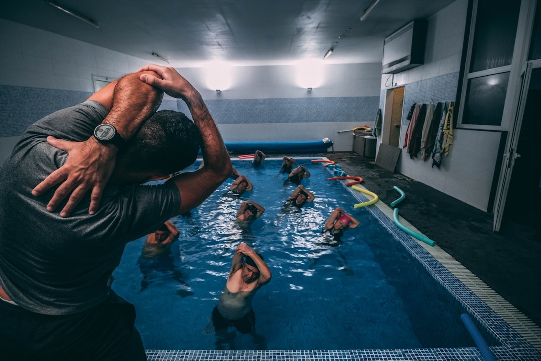 En grupp deltagare i en inomhusbassäng utför synkroniserade övningar under vattnet ledda av en instruktör, vilket visar ett strukturerat träningsprogram för vattenfitness.